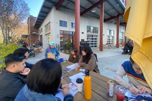 Adults sitting at a table in discussion. Some are talking, some listening, and some taking notes. There are snacks and drinks present.