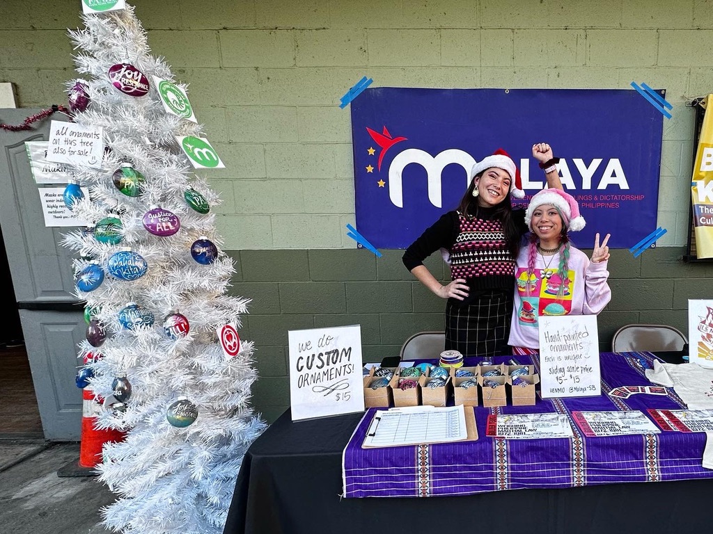 2 malaya members Alyssa and Kat posing at their table. There's a christmas tree adorned with hand painted ornaments